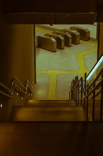 View from the top of a staircase leading down into an underground parking or transportation area. The staircase has metal handrails on both sides, and the steps are wooden with a dark finish. At the b