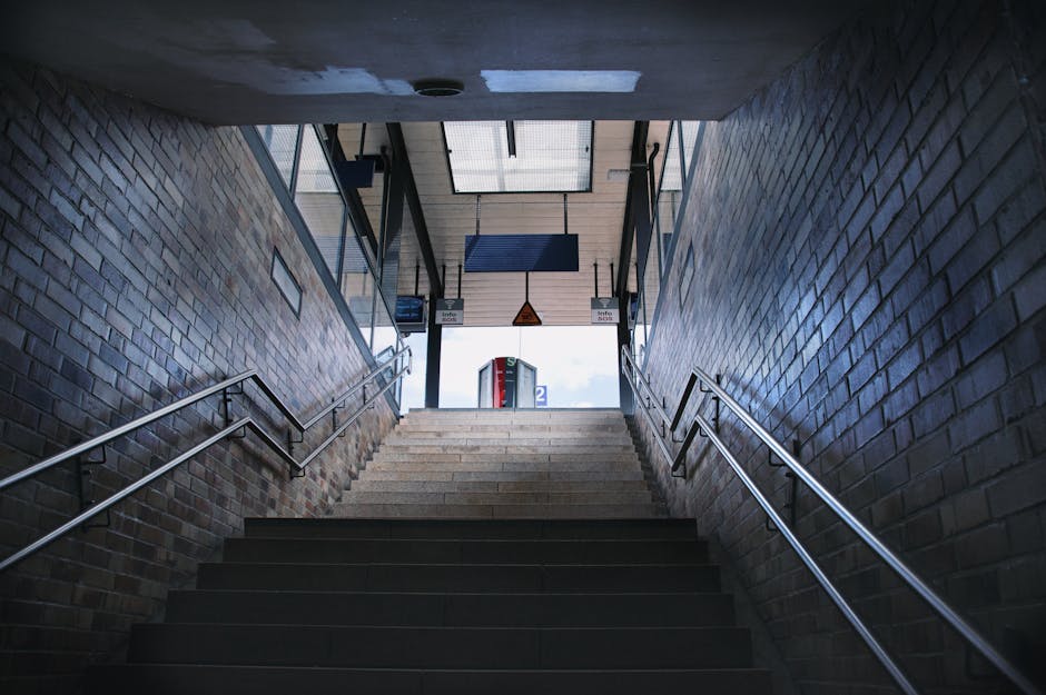 View from the top of a staircase leading down into an underground parking or transportation area. The staircase has metal handrails on both sides, and the steps are wooden with a dark finish. At the b