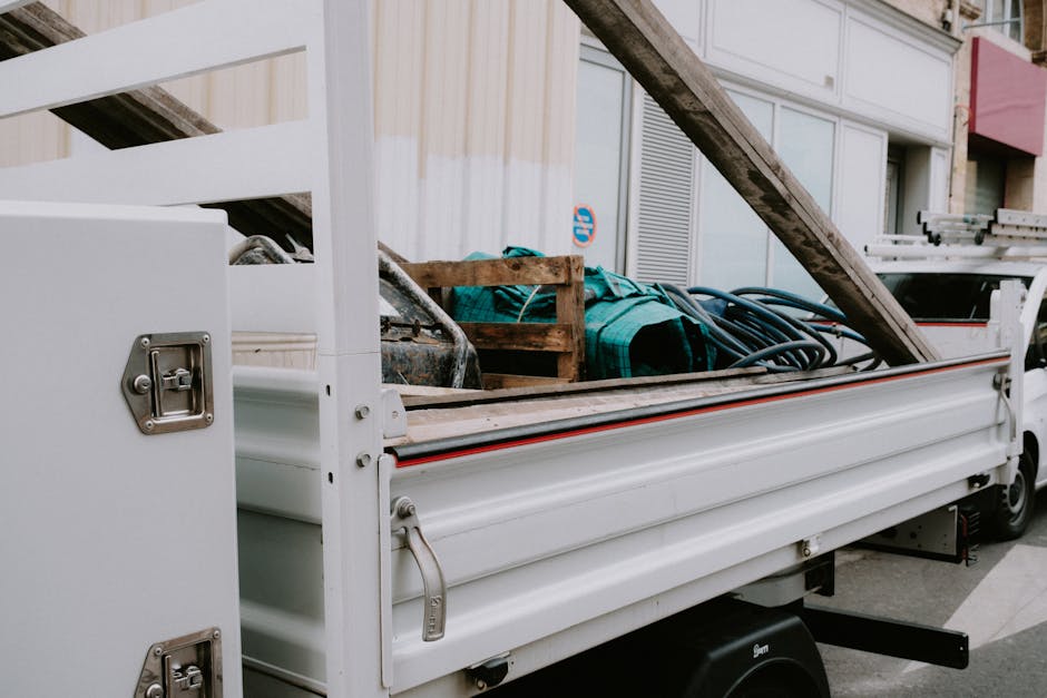 A collection of discarded household items and debris, including a metal-framed bed slatted frame, broken wooden planks, a damaged dresser with its drawer missing, and various pieces of cardboard and p