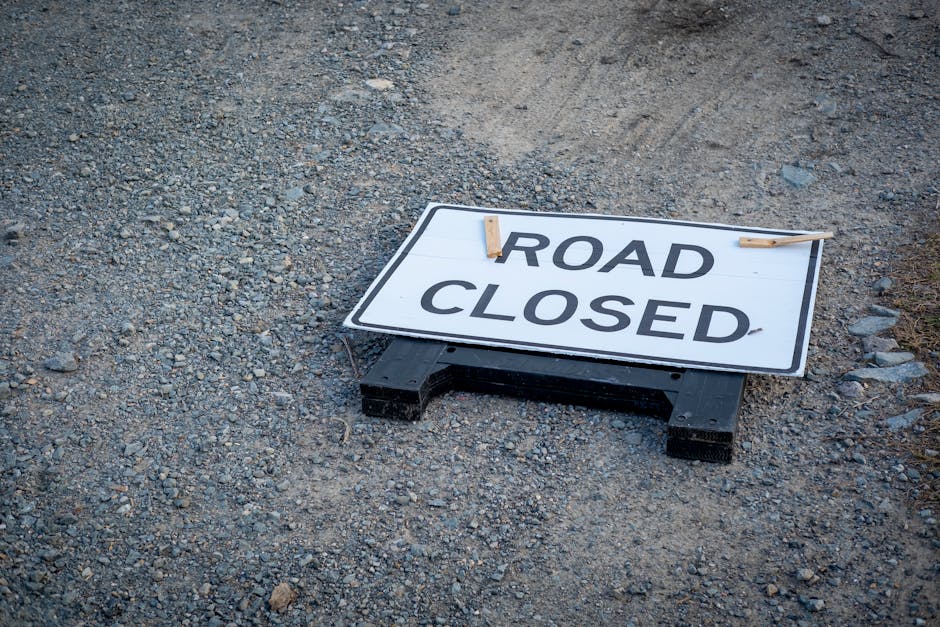 A snow-covered outdoor scene featuring a closed road with a barrier composed of horizontal red and white striped bars. Behind the barrier are three amber-colored warning lights attached to the top of