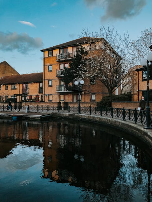 A multi-storey brick residential building with small balconies is situated by a waterway, with a paved walkway and black metal railing along the edge. Several trees, some with bare branches, are posit